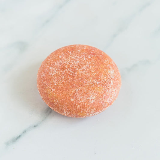 A round, pink sulfate free shampoo bar shown on a marble countertop.