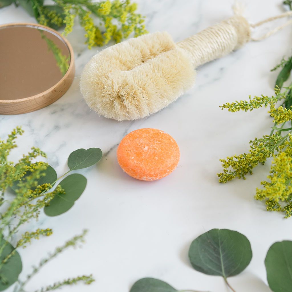 A shampoo bar on a marble background next to green leaves, flowers and a brush