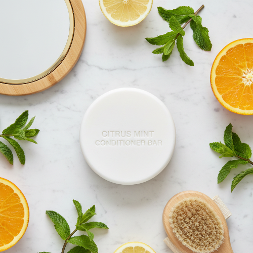 An energizing flat lay of a natural Citrus Mint Conditioner Bar on marble, featuring bright citrus fruit, fresh herbs, a wooden hand mirror, and a bath brush to showcase its refreshing and eco-friendly ingredients.