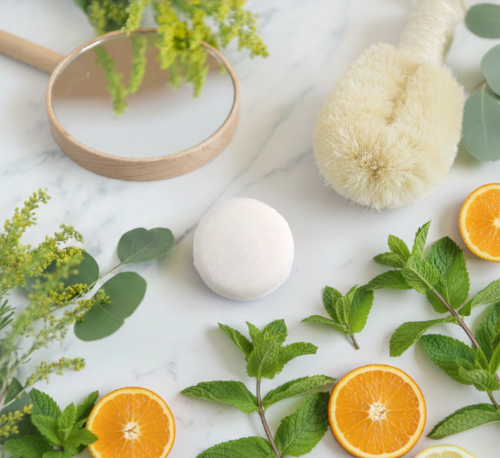 A professional product photograph of a bright, round Citrus Mint Shampoo Bar on a white marble surface, styled with fresh orange slices, peppermint leaves, and eco-friendly wooden bath accessories.