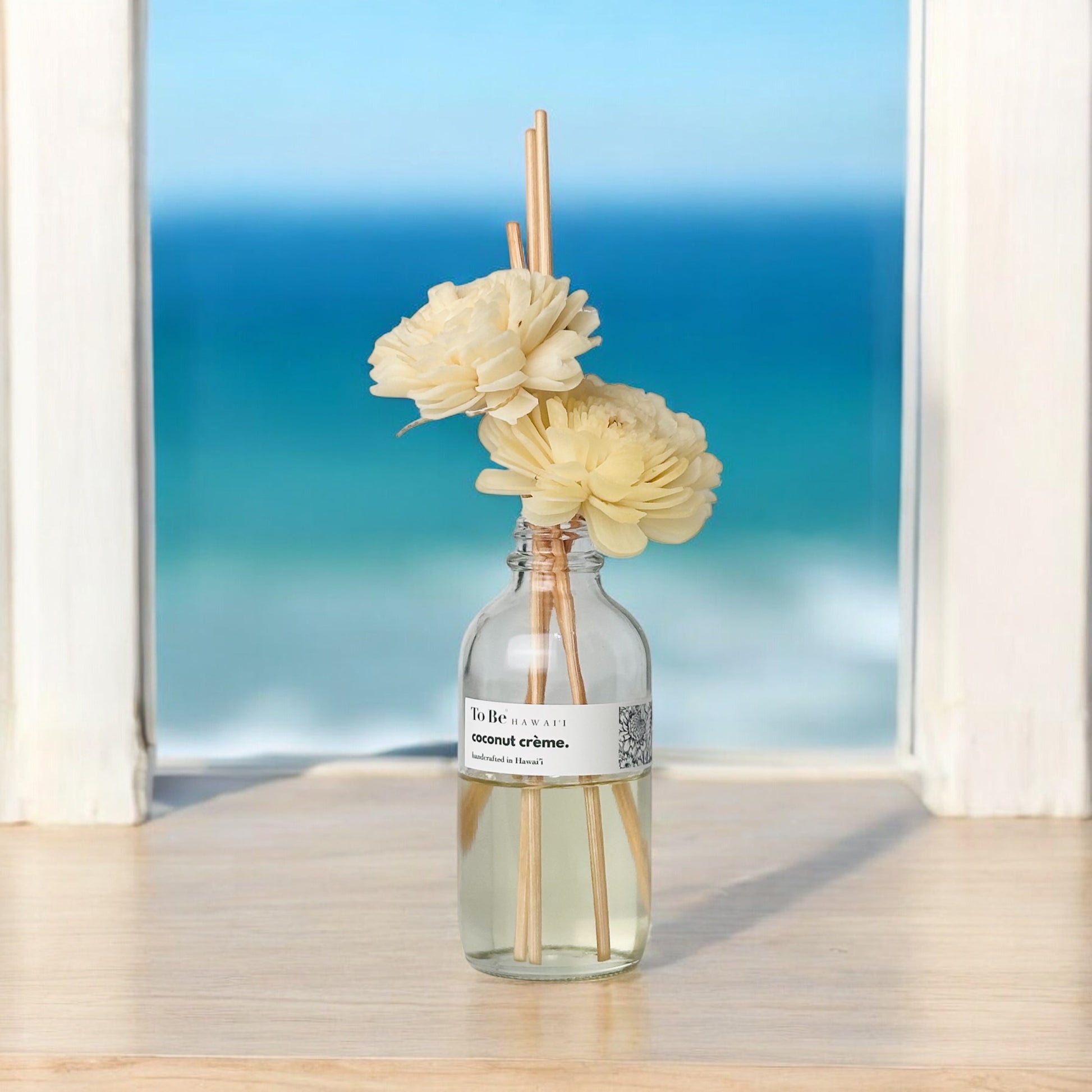 Reed diffuser with flowers on a table in front of a window with a blue ocean view