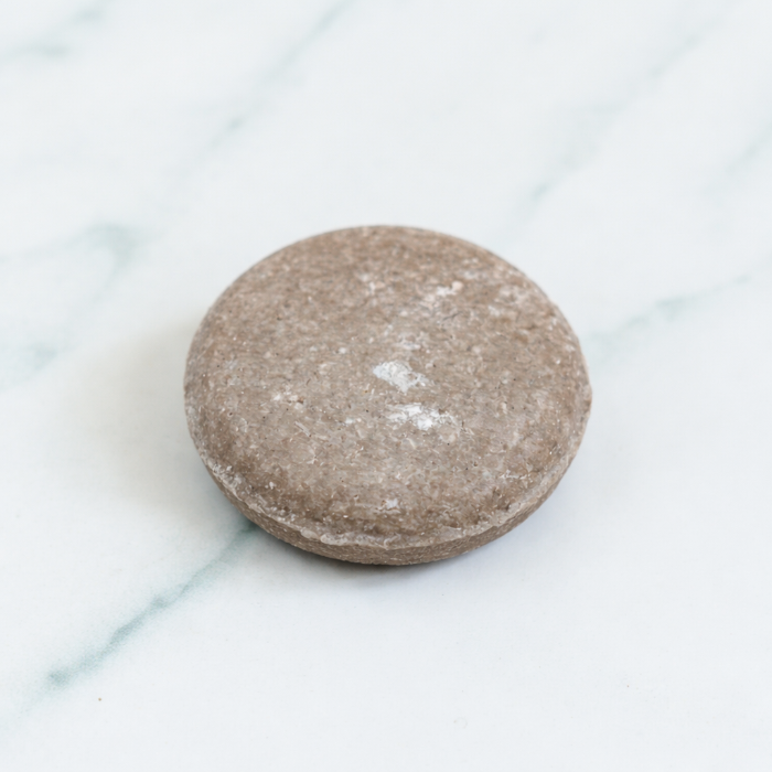 A close-up, top-down studio photograph of a textured, light-purple Rosemary Shampoo Bar centered on a clean white marble surface, surrounded by fresh green rosemary sprigs and botanical leaves.
