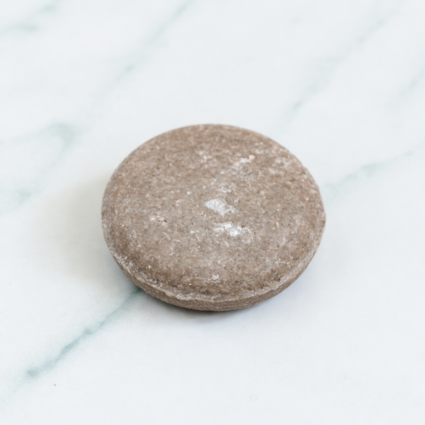 A professional close-up of a textured, earth-toned Rosemary Shampoo Bar. The bar is shown on a clean marble background, accompanied by fresh rosemary leaves and a wooden handle brush to emphasize its natural, stimulating ingredients.