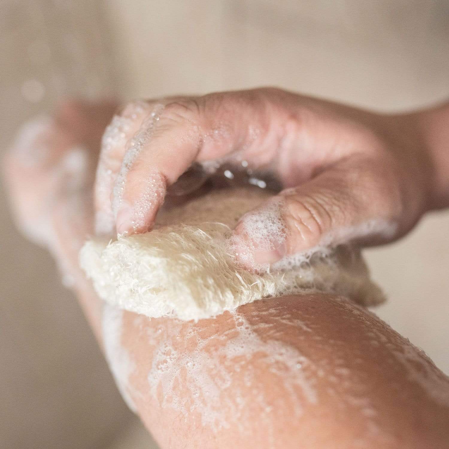 Close-up of a person using a natural loofah sponge to lather a bar of soap, showing the fibrous texture and soapy suds against skin.