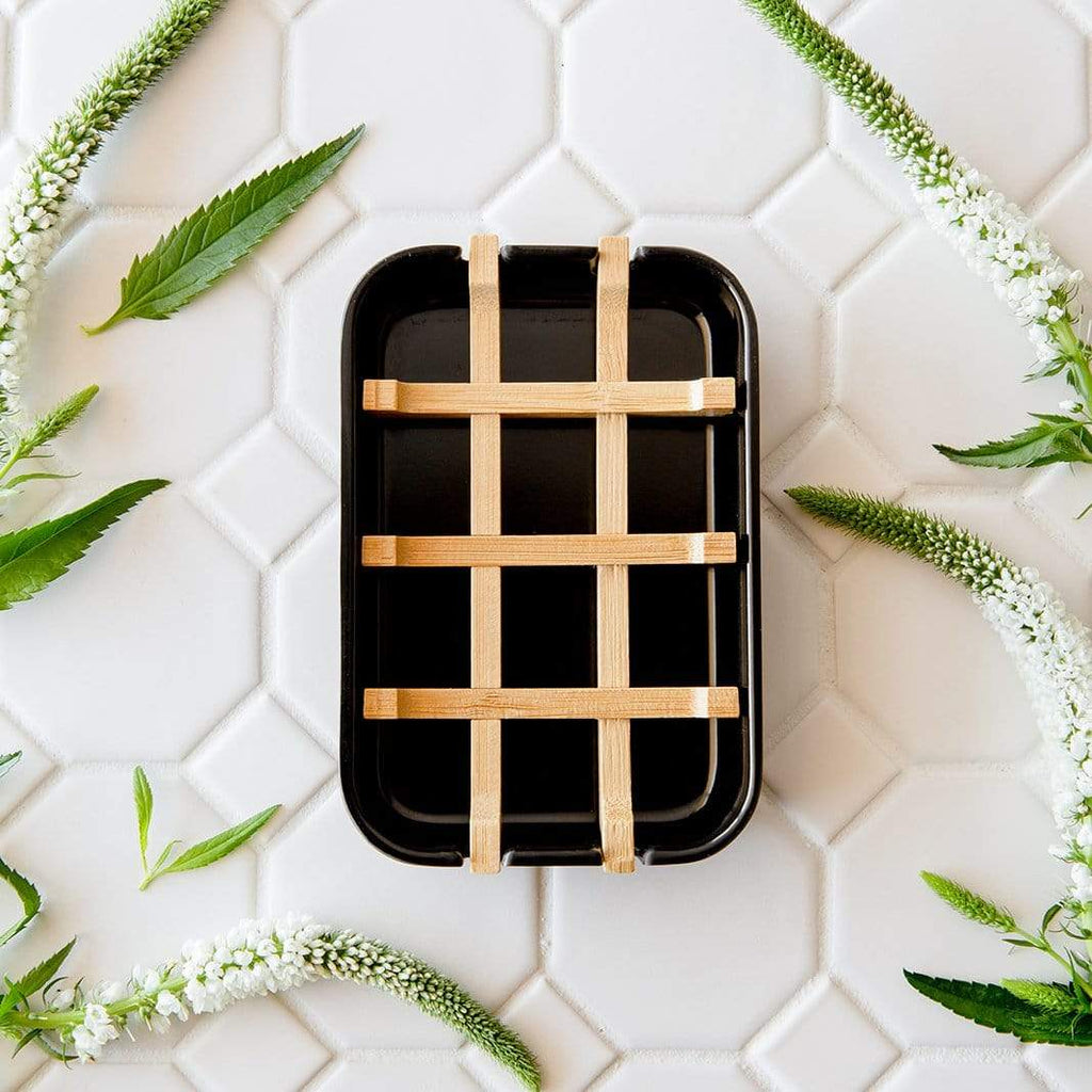 Black rectangular bar soap hold with woven wooden handles on a white tiled background with green leaves.