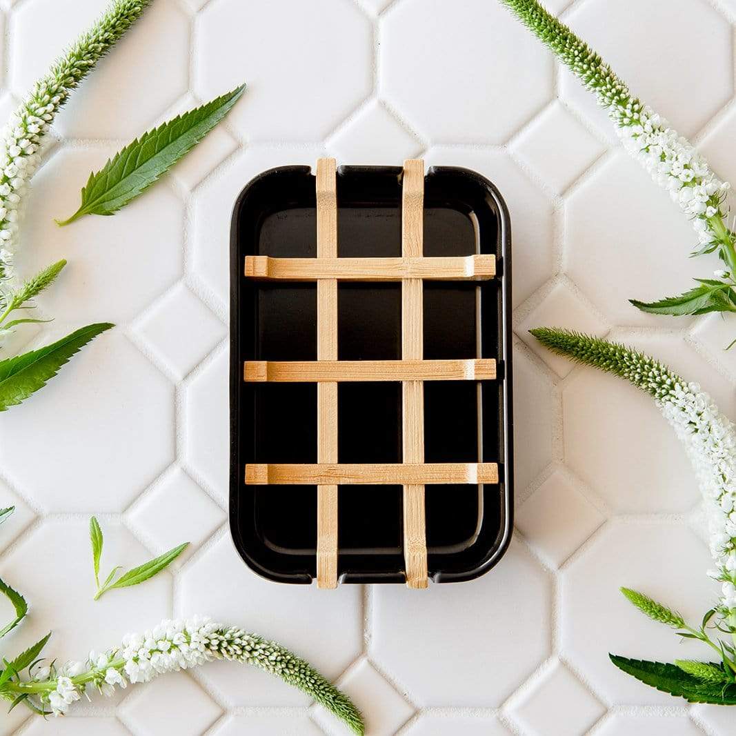 Black rectangular bar soap hold with woven wooden handles on a white tiled background with green leaves.