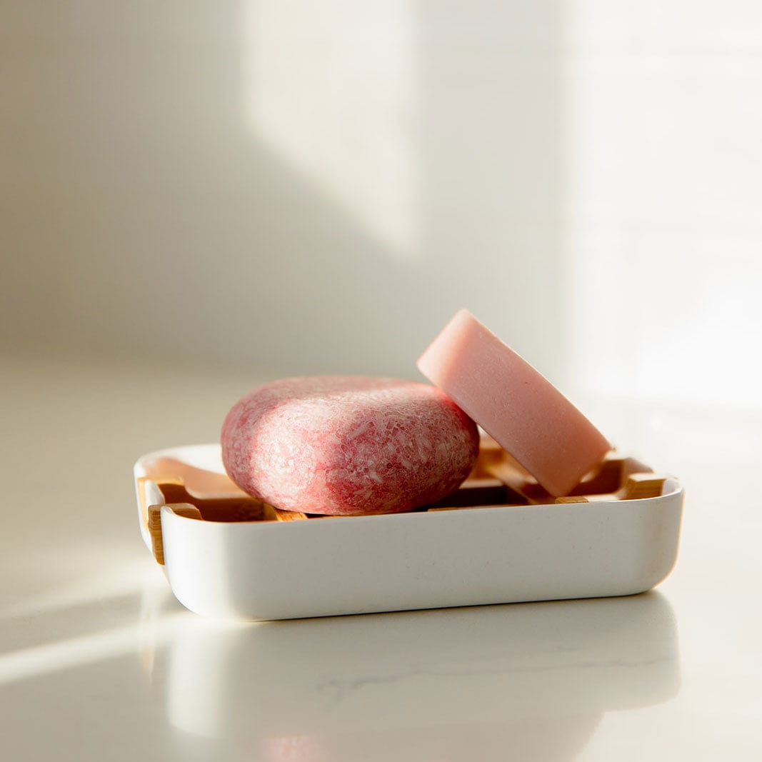 Pink shampoo and conditioner bars on a white soap dish with a soft light background