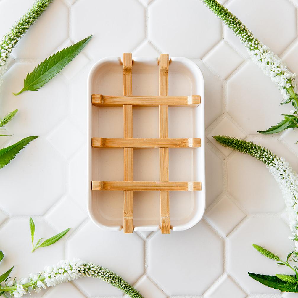 A white bar soap holder with a natural bamboo grid insert, displayed on a white hexagonal tile background with green botanical accents.
