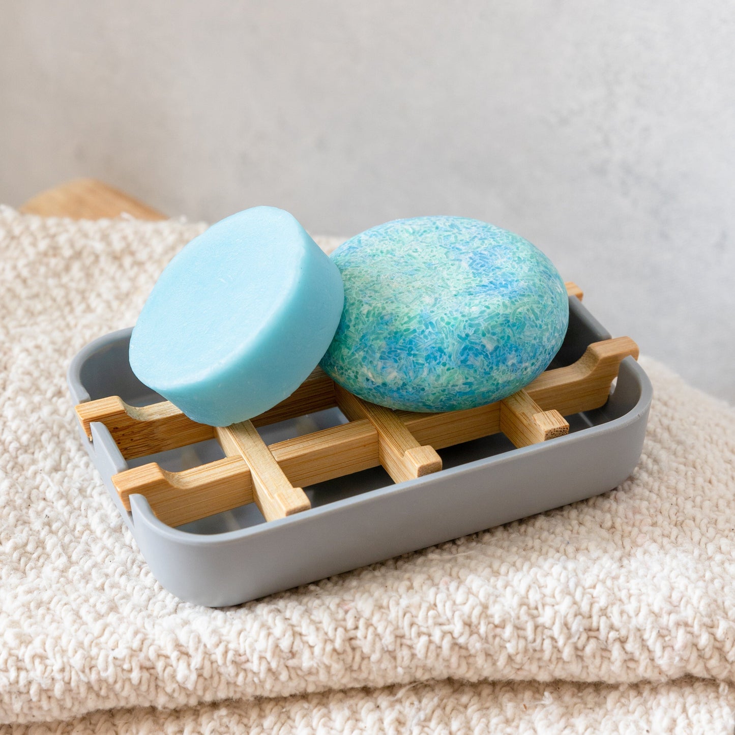 Two blue shampoo bar and conditioner on a gray soap dish with wooden support on a beige towel.