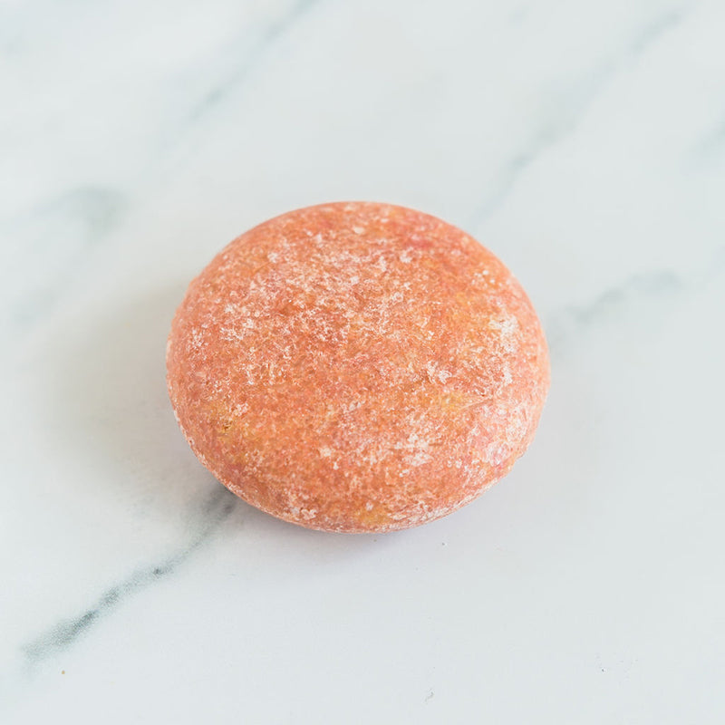 A round, pink sulfate free shampoo bar shown on a marble countertop.