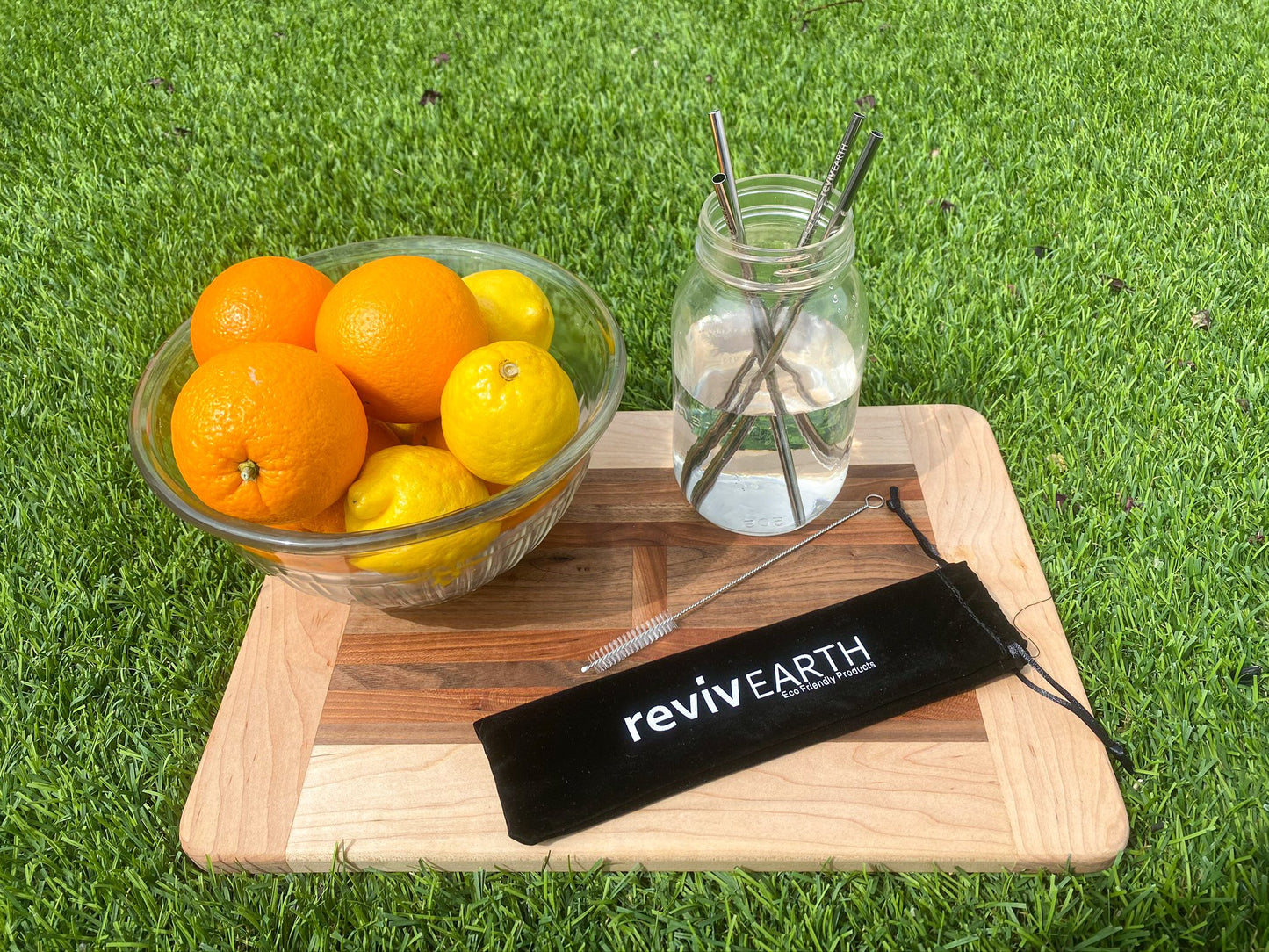 A straw stainless steel set of four inside a glass of water, displayed on a wooden cutting board outdoors with a bowl of citrus and a black revivEARTH carrying pouch.