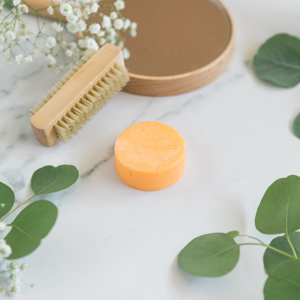 An orange, round best bar conditioner centered on a marble surface, styled with a wooden hairbrush and fresh eucalyptus leaves for an eco-friendly look.
