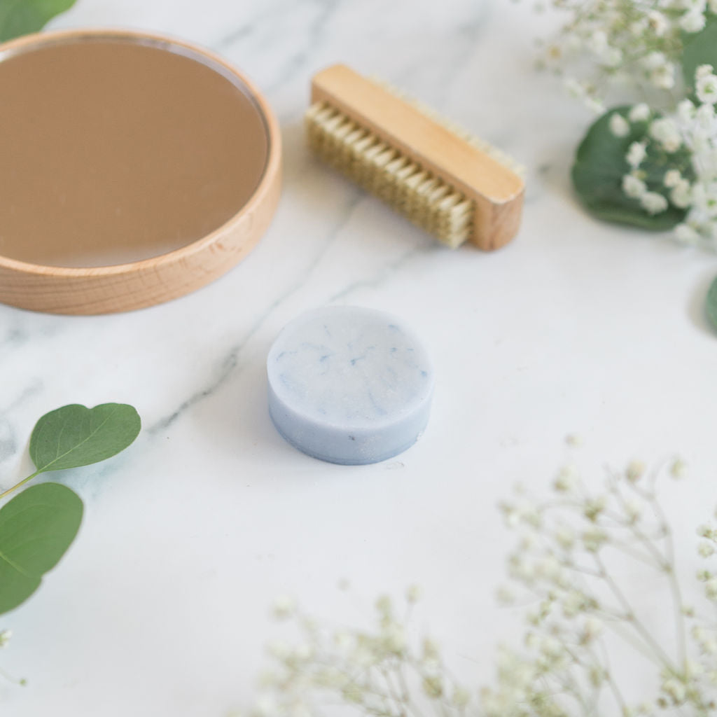 A blue conditioner bar soap for men centered on a marble surface with a wooden nail brush and fresh eucalyptus leaves.