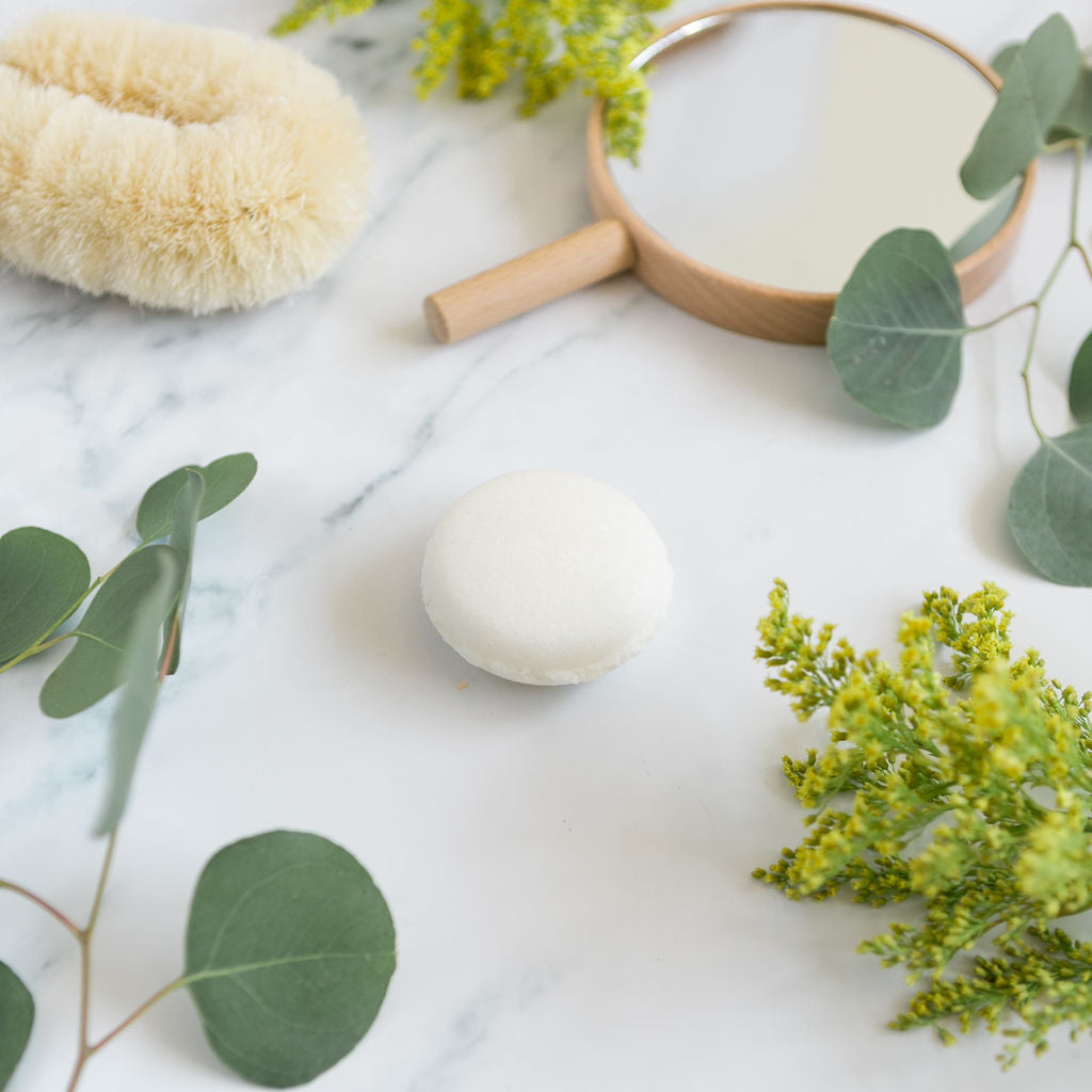 A white, round shampoo that are sulfate free bar displayed on a marble surface with a natural sisal brush, green eucalyptus leaves, and yellow botanicals.