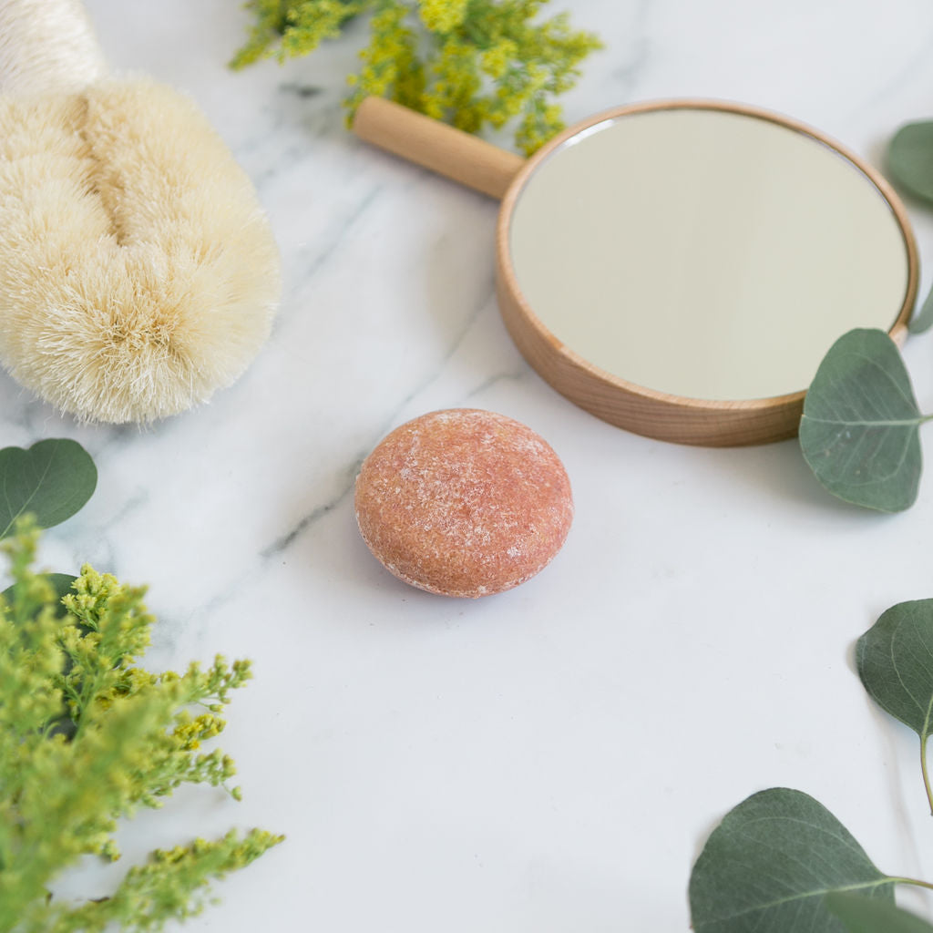 A round, pink sulfate free shampoo bar shown on a marble countertop alongside a mirror and fresh eucalyptus leaves.