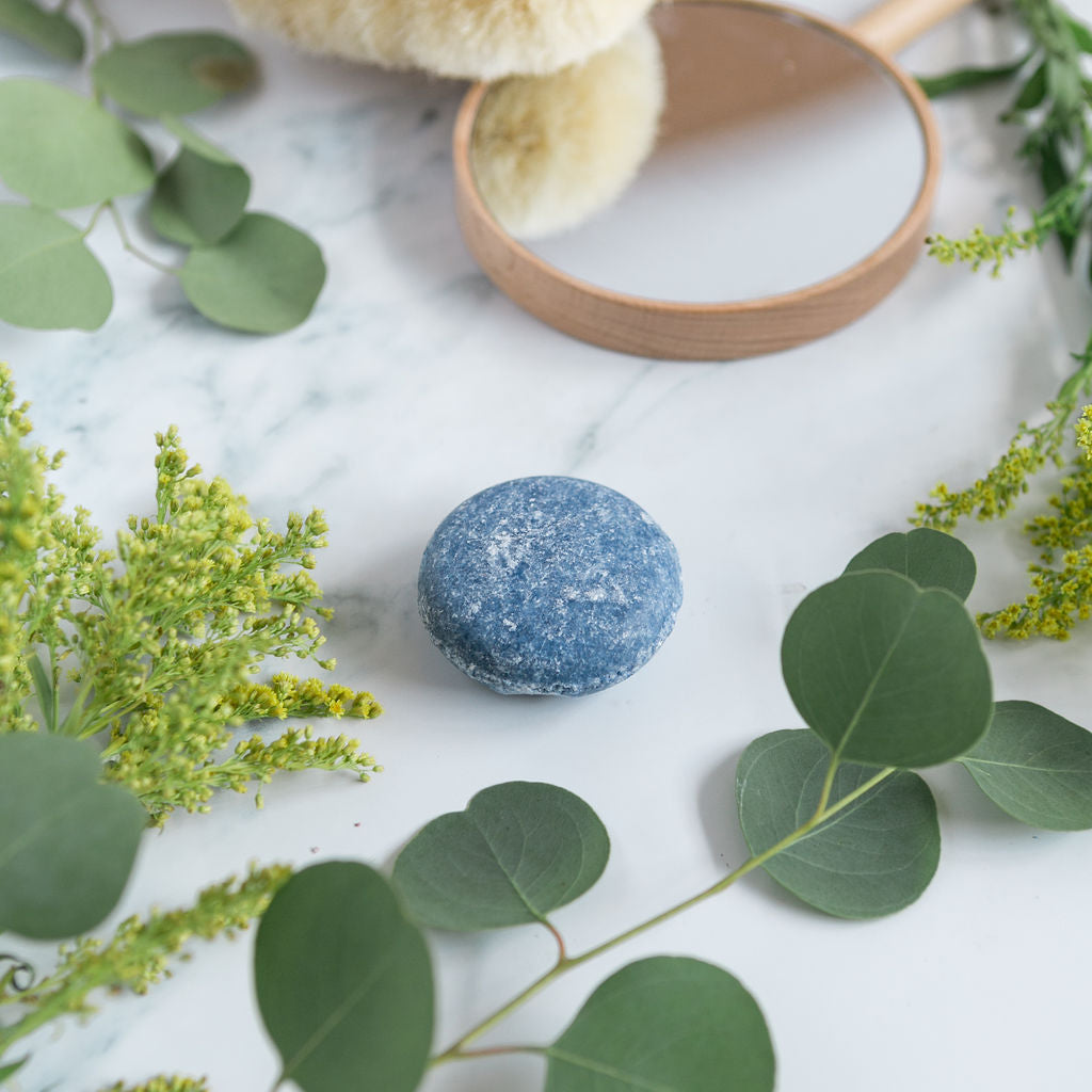A blue no plastic shampoo bar for men centered on a white marble surface, surrounded by fresh eucalyptus leaves and a wooden brush.